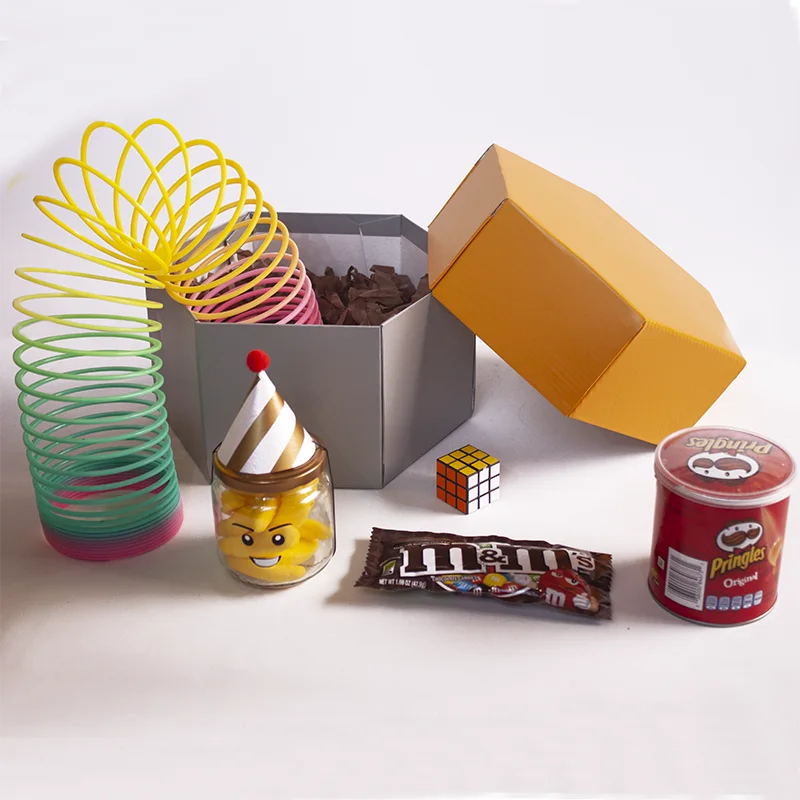 Caja en forma hexagonal, con un frasco de gomas, mini cubo rubik, papas Pringles y resorte de plástico de colores.